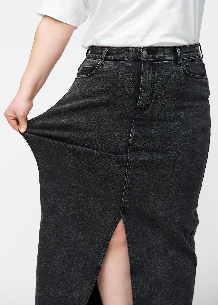 Plus-size model strækker den lange, mørkegrå denimnederdel i farven Shadows ved hoften for at vise materialets stretch.