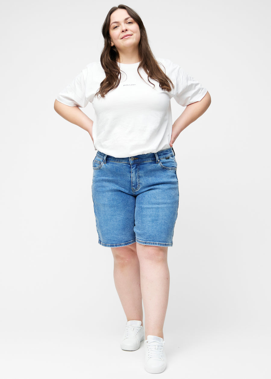 Fuldt billede af plus-size model i løse, klassisk blå denim shorts i farven Rivers, der går til lige over knæet.