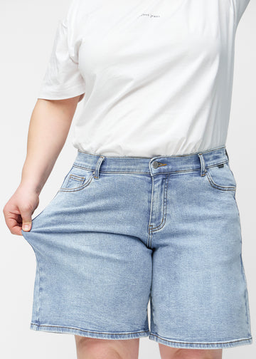 Plus-Size-Model dehnt die lockeren, hellblauen Denim-Shorts in der Farbe Waves an der Hüfte, um den Stretch des Materials zu zeigen.