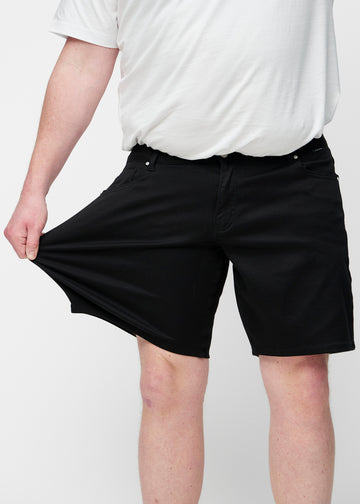 Plus-Size-Modell dehnt schwarze Shorts für Herren in der Farbe Ravens am Oberschenkel, um den Stretch des Materials zu zeigen.