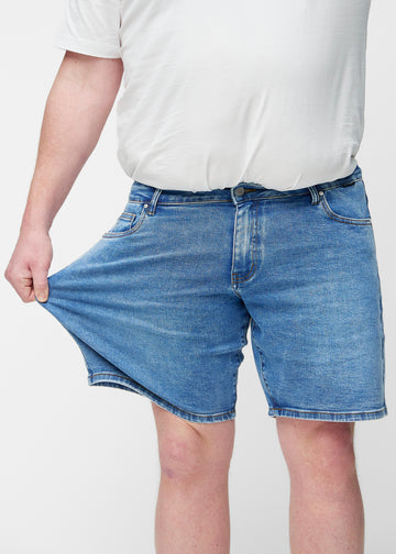Plus-Size-Modell dehnt blaue Denim-Shorts für Herren in der Farbe Rivers am Oberschenkel, um den Stretch des Materials zu zeigen.