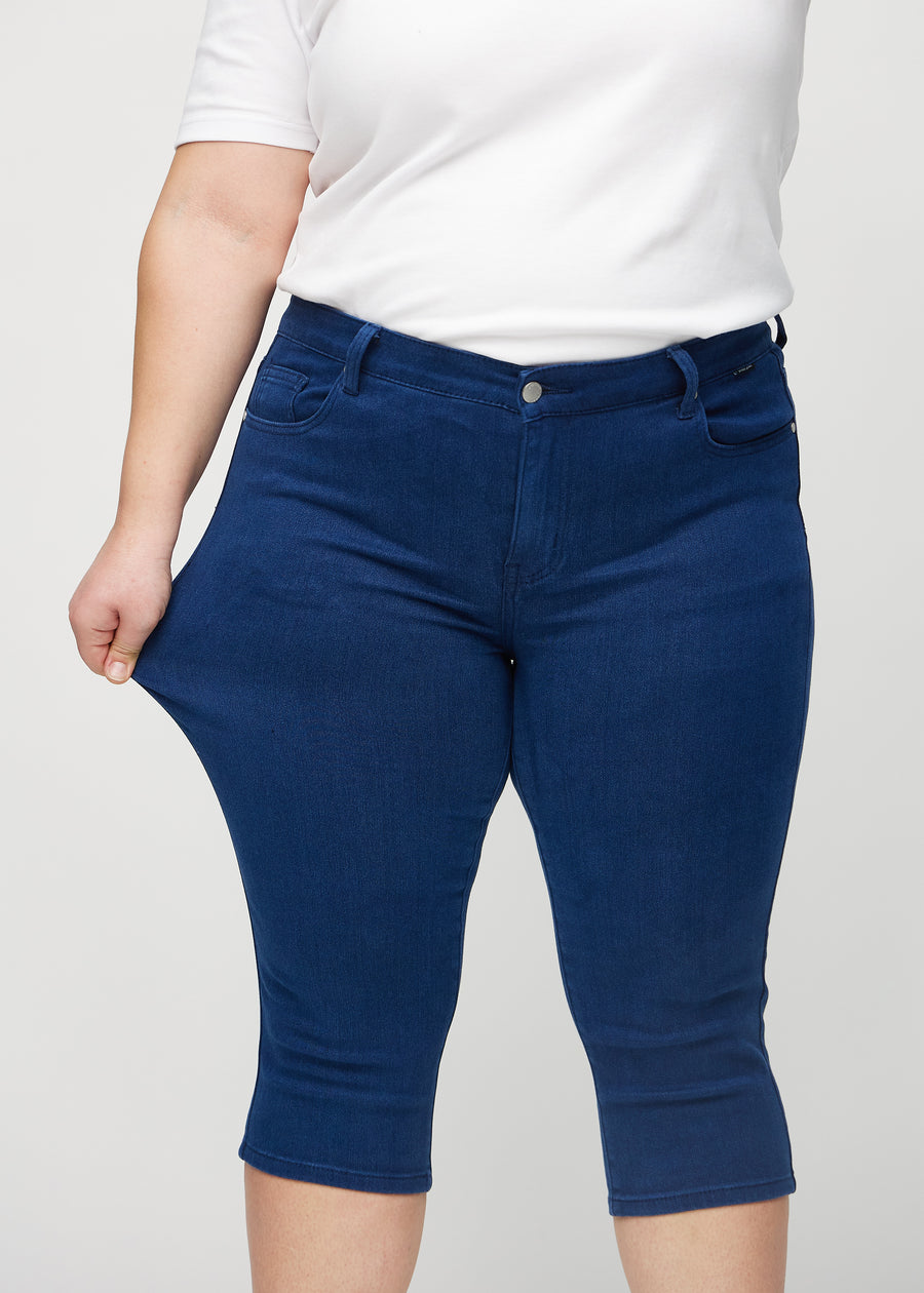 Skinny-Capris für Damen in dunkelblauem Jeansstoff, Farbe Royals. Ein Plus-Size-Model dehnt den Stoff, um den Stretch zu zeigen.