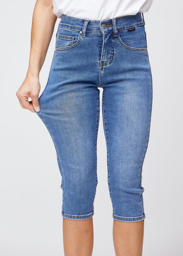 Slim-Capris für Damen aus blauem Denim, Farbe Rivers. Ein Model dehnt den Stoff am Oberschenkel, um die Flexibilität zu zeigen.