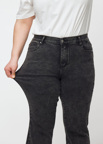 Dunkelgraue Mid-Rise-Bootcut-Jeans in der Farbe Shadows. Ein Plus-Size-Model zieht am Jeansstoff am Oberschenkel, um den Stretch zu zeigen.