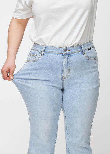 Hellblaue Mid-Rise-Bootcut-Jeans in der Farbe Waves. Ein Plus-Size-Model zieht am Jeansstoff am Oberschenkel, um den Stretch zu zeigen.