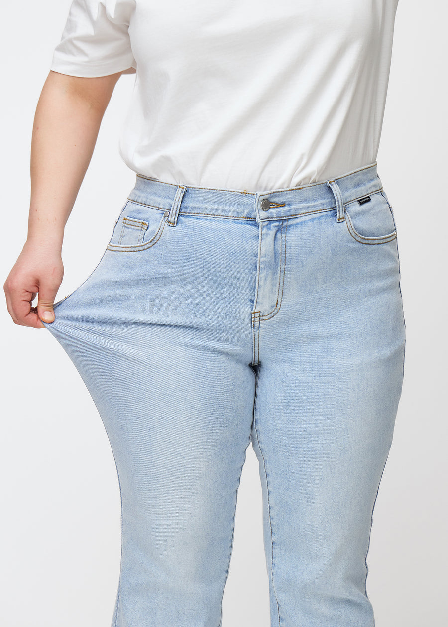 Hellblaue Mid-Rise-Bootcut-Jeans in der Farbe Waves. Ein Plus-Size-Model zieht am Jeansstoff am Oberschenkel, um den Stretch zu zeigen.