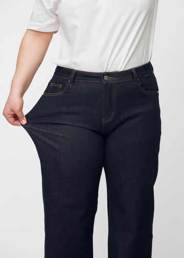 Ein Plus-Size-Model dehnt die tief dunkelblaue Denim jeans für Damen in der Farbe Midnights am Oberschenkel, um die Dehnbarkeit des Materials zu zeigen.