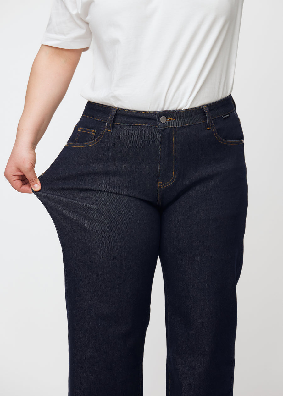 Ein Plus-Size-Model dehnt die tief dunkelblaue Denim jeans für Damen in der Farbe Midnights am Oberschenkel, um die Dehnbarkeit des Materials zu zeigen.