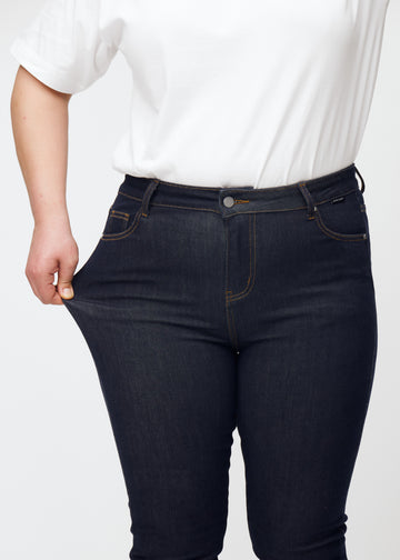 Ein Plus-Size-Model dehnt die tief dunkelblaue Denim jeans für Damen in der Farbe Midnights am Oberschenkel, um die Elastizität des Materials zu zeigen.
