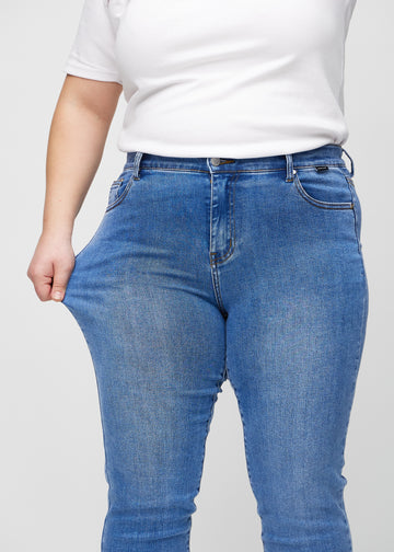 Slim-Jeans für Damen in Blau, Farbe Rivers. Ein Plus-Size-Model zieht am Jeansstoff am Oberschenkel, um den Stretch zu zeigen.