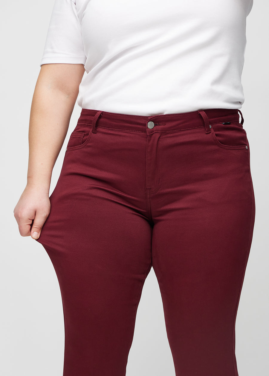 Slim-Jeans für Damen in Dunkelrot, Farbe Santas. Ein Plus-Size-Model zieht am Jeansstoff am Oberschenkel, um den Stretch zu zeigen.