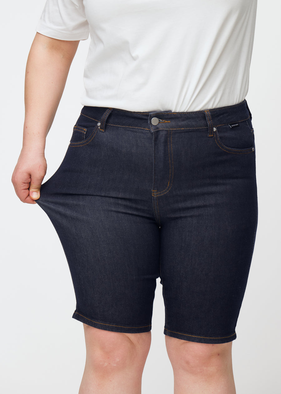 Plus-Size-Model dehnt tief dunkelblaue Denim-Shorts für Damen in der Farbe Midnights am Oberschenkel, um den Stretch zu zeigen.