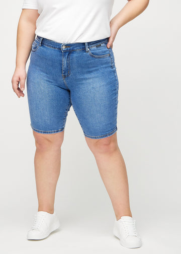Denim skinny middle shorts, Modellname Rivers, die eng am Oberschenkel anliegen und direkt über dem Knie enden, an einem Plus-Size-Model von vorne gesehen.