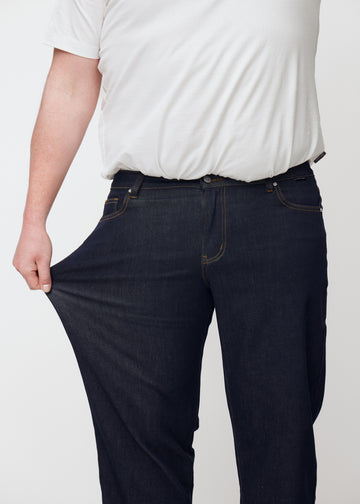 Das Plus-Size-Model dehnt die tief dunkelblaue Denim jeans für Herren in der Farbe Midnights am Oberschenkel, um die Stretchqualität des Materials zu zeigen.