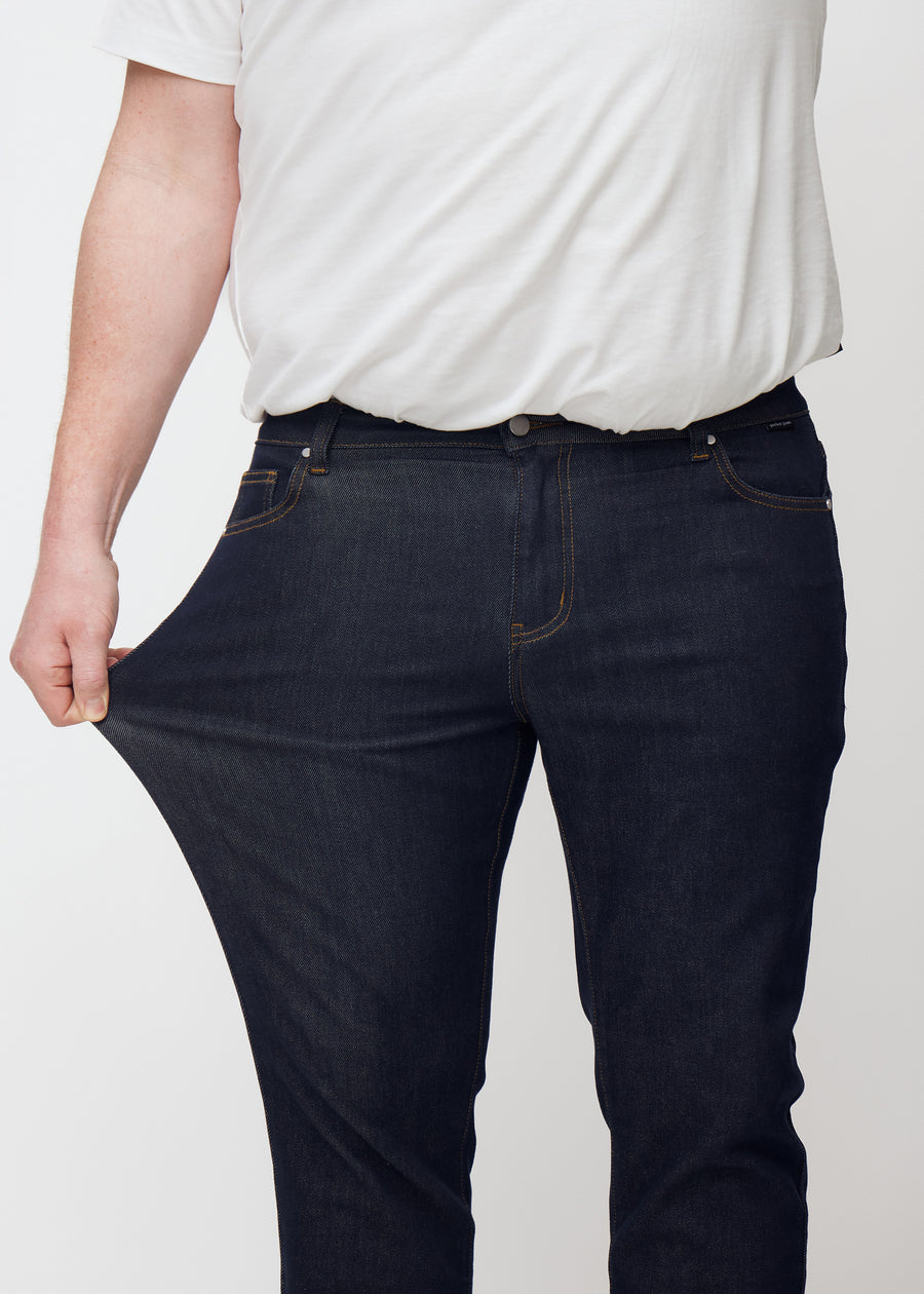 Das Plus-Size-Model dehnt die tief dunkelblaue Denim jeans für Herren in der Farbe Midnights am Oberschenkel, um die Dehnbarkeit des Materials zu zeigen.