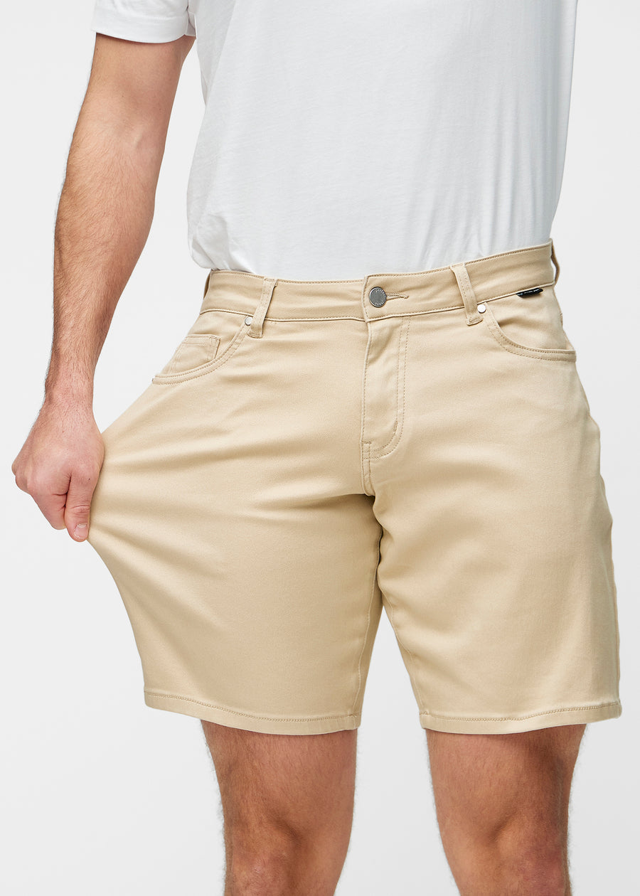 Modell dehnt beige Shorts für Herren in der Farbe Gazelles am Oberschenkel, um den Stretch des Materials zu zeigen.