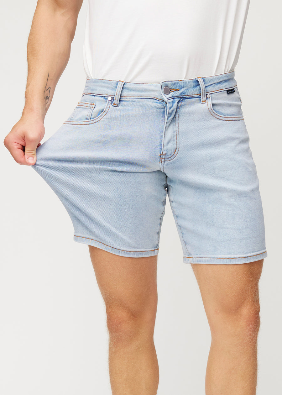 Das Model dehnt hellblaue Denim-Shorts für Herren in der Farbe Waves am Oberschenkel, um den Stretch des Materials zu zeigen.